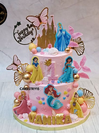 A grand two-tier fairytale princess cake for a first birthday. This pineapple cake is adorned with figures of multiple Disney princesses like Snow White, Ariel, and Cinderella, against a pink castle backdrop.