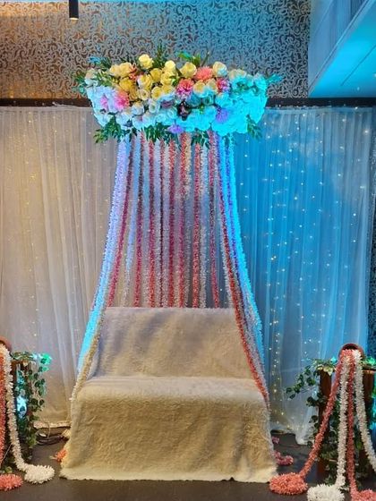 A canopy-style seating arrangement for a Mehndi function. This design features a floral chandelier with hanging flower strings in pink and white over the main seat.