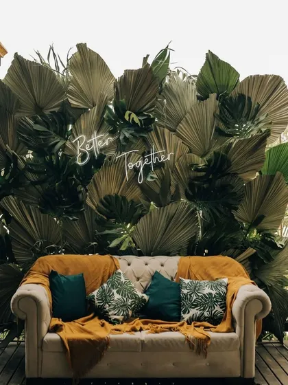 A chic and modern boho-themed photo corner with a 'Better Together' neon sign. The backdrop of large dried palm leaves and a comfortable tufted sofa creates a stylish, earthy look.
