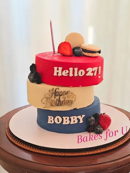A unique, modern three-tier cake for a 27th birthday. Each tier has a different color and message, creating a stacked, minimalist design topped with fresh berries and macarons.