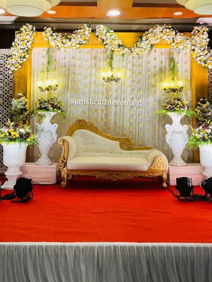 A full view of a classic engagement stage setup. It features a white and gold chaise, decorative urns, and a backdrop of white drapes framed by yellow and white floral decor.