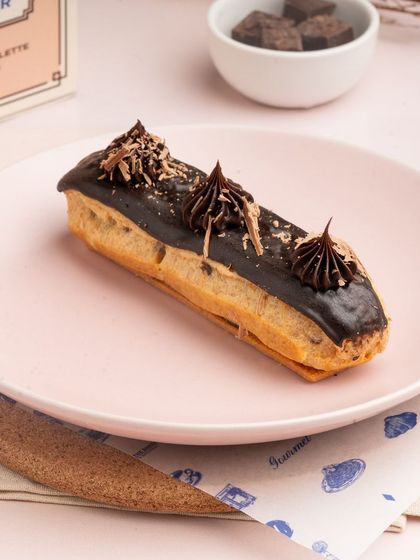 A perfectly crafted chocolate eclair, sitting on a pink plate, ready to be enjoyed. The chocolate shavings on top add a touch of elegance.