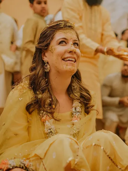 The bride's face, smeared with turmeric, is lit up with a beautiful, happy smile. This close-up captures the pure, unscripted joy of the Haldi ceremony.