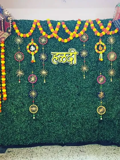 A clean and traditional Haldi backdrop for home functions. The green wall is decorated with marigold garlands and colorful hangings, creating a perfect photo spot.