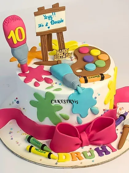 A colorful single-tier artist-themed cake for a 10th birthday. The cake is decorated with a fondant easel, paint palette, brushes, and crayons, celebrating a decade of creativity.