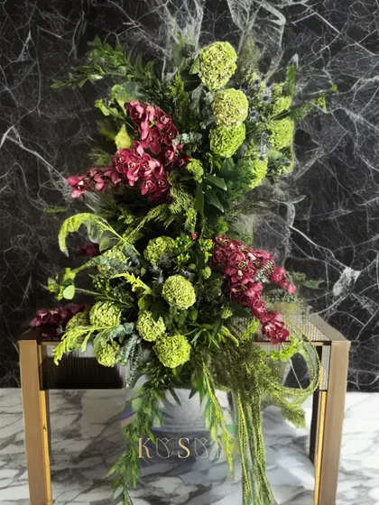 A dancing ballerina for your living room console. This tall, elegant flower installation uses a unique vertical structure to bring life and movement to an interior space, featuring lush greens and pops of deep magenta.