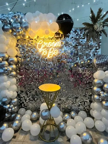 Celebrate the groom with a modern and stylish party setup. This "Groom to Be" decoration features a dazzling silver shimmer wall, a chic neon sign, and a sophisticated balloon arrangement in black, silver, and white, perfect for a bachelor party.