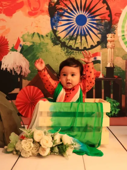 A baby joyfully raises his hands during an Independence Day photoshoot. He is peeking out of a crate draped with the Indian tricolor, set against a vibrant, patriotic-themed background.
