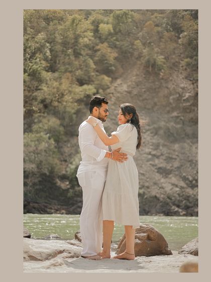 A full-length portrait of the couple in white, standing by the Ganga, framed to feel like a magazine cover.