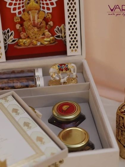 This elegant white invitation box features a laser-cut Ganesha centerpiece. The compartments hold gourmet treats in test tubes and jars, alongside a miniature elephant idol, blending sacred motifs with modern gifting trends.