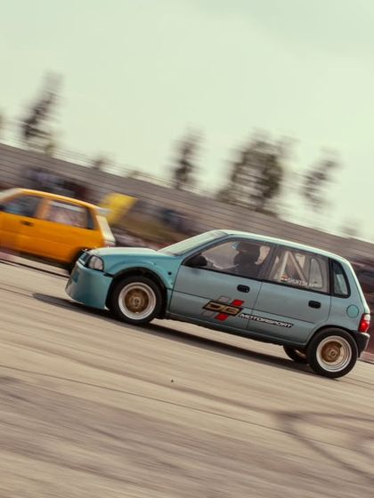 Two modified hatchbacks, a blue Chevy and a yellow Suzuki, in a close race. The competition in these classes is just as fierce as in the supercar categories.