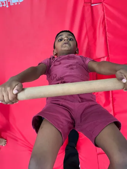 A young boy holds a pull-up on a wooden bar, showing his determination and growing strength. This simple exercise is incredibly effective for building the upper body muscles needed for gymnastics.