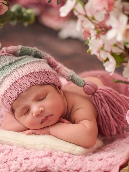 Framed by soft pink cherry blossoms, a sleeping newborn wearing a striped tassel hat rests on a pink blanket, capturing the gentle beauty of spring.