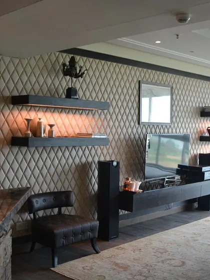 This living room is a study in sophisticated comfort. The feature wall combines quilted leather panels with floating shelves, creating a textured backdrop for the entertainment unit. The reclaimed wood bar adds a rustic yet luxurious element to the space.