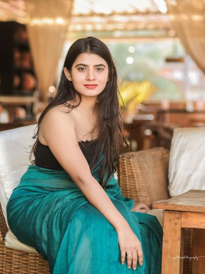 A modern take on saree styling. The model wears a teal saree paired with a contemporary black top, shot in a cafe with earthy tones. This shows how traditional wear can be styled for a modern, chic look.