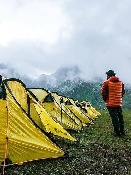 A line of tents ready for a group of adventurers, facing a majestic, cloud-covered mountain range. This is a moment of pause, taking in the scale of the Himalayas before settling in for the night.