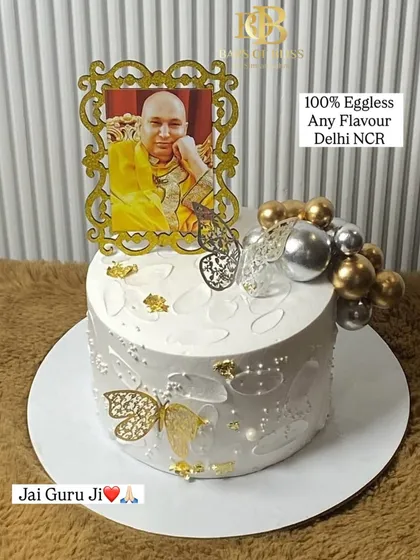 A cake made with devotion for a Guruji celebration. This elegant white cake is decorated with a photo of Guruji in a golden frame, delicate butterflies, and gold and silver spheres.