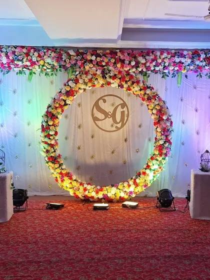 A classic and elegant engagement stage backdrop. The focal point is a large, circular ring completely covered in multi-colored flowers, with a custom monogram of the couple's initials placed in the center for a personal touch.