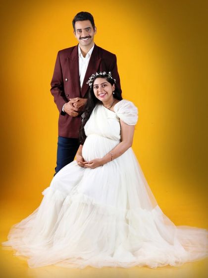 A classic studio portrait of an expecting couple. The mom-to-be wears an elegant white gown with puffy sleeves, which stands out beautifully against the solid yellow backdrop.