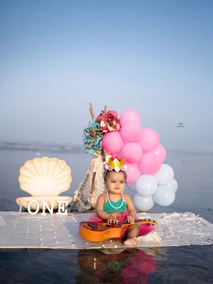 A little girl dressed as a mermaid for her first birthday beach photoshoot. The setup includes a seashell lamp, a ukulele, and pink balloons, creating a magical, themed celebration.