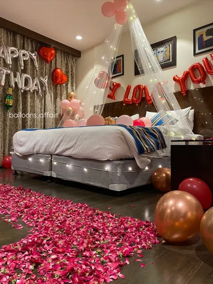I designed this hotel room for a truly romantic birthday surprise. It features a beautiful rose petal pathway leading to a bed adorned with a delicate canopy and fairy lights, complemented by rose gold and red balloons.
