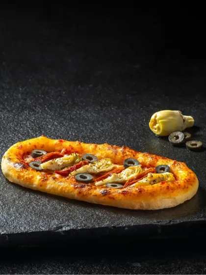 An oval-shaped flatbread pizza with artichokes and olives, styled minimally on a dark slate to emphasize its unique shape and toppings.