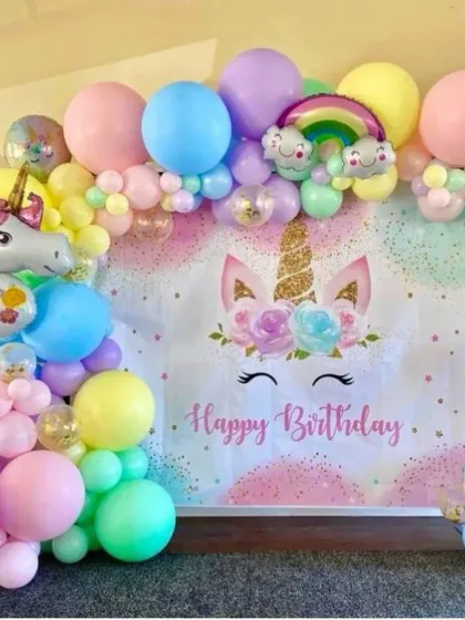 An enchanting unicorn-themed first birthday party. A custom backdrop is framed by a pastel rainbow balloon garland, complete with a large unicorn foil balloon and a number "1" balloon.