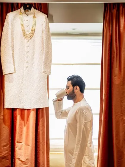 A creative shot of the groom enjoying a quiet cup of coffee before getting into his sherwani, which hangs in the background. A moment of calm before the celebration begins.