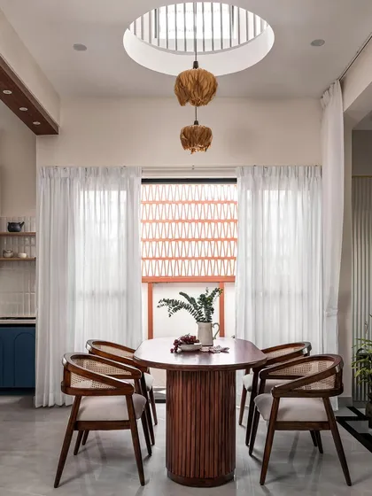 This dining area is defined by a double-height cutout in the ceiling, allowing natural light to flood the space. A cluster of pendant lights hangs through the opening, connecting the ground floor to the level above and creating a dramatic focal point.