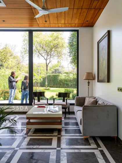 This seating area in 'The Green Axis' is positioned to take full advantage of the garden view through a large picture window. The wood ceiling adds warmth, contrasting with the cool black-and-white marble floor.