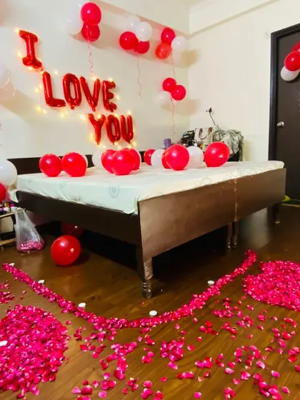A simple yet impactful romantic gesture. This room features 'I LOVE YOU' foil balloons lit by fairy lights, a floor covered in a pathway of rose petals, and red and white balloons scattered around.