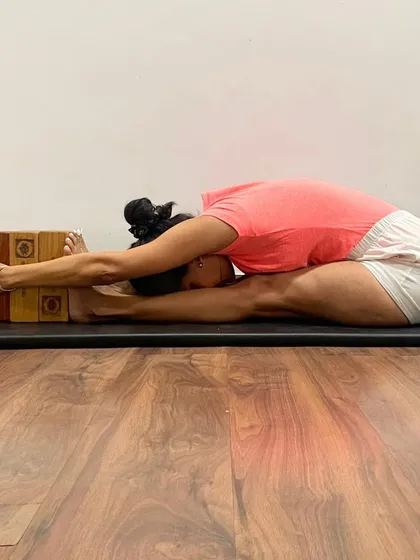 Janu Sirsasana (Head-to-Knee Pose) is demonstrated with blocks supporting the arms. This allows the torso to extend forward fully, creating a deep stretch for the hamstrings and a gentle compression of the abdominal organs.