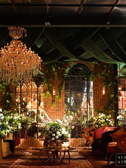 A wide view of the glass house reception, showing the dramatic green fabric draped from the ceiling, the warm glow of the chandeliers, and the inviting lounge areas.