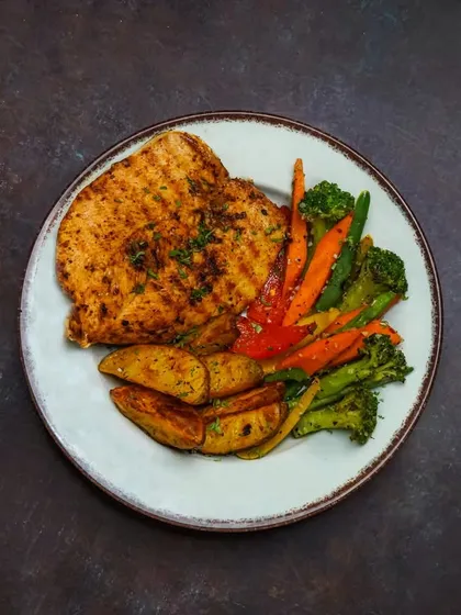 A grilled chicken platter served with roasted vegetables and potato wedges. The top-down angle on a dark, rustic surface gives the image a warm and hearty feel.