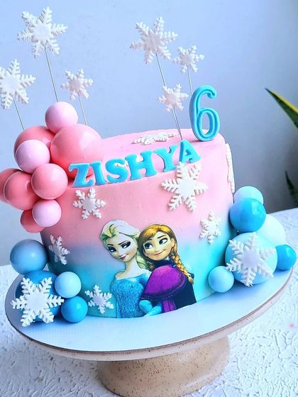 A Frozen cake with a pink and blue ombre effect, decorated with edible snowflakes and chocolate spheres.