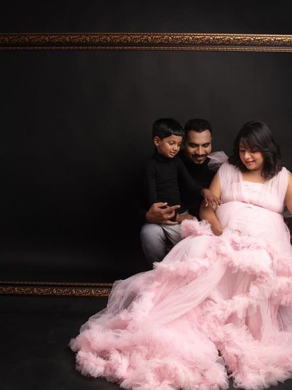A creative family portrait using a large, empty frame as a prop. The mom-to-be's soft pink gown provides a beautiful pop of color against the dark background.