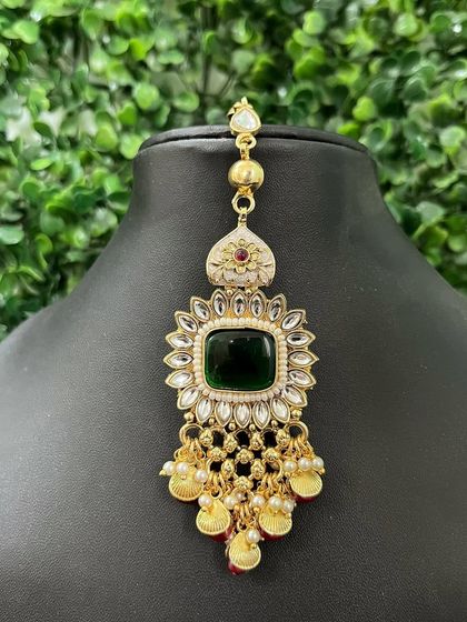 A statement maang tikka with a large square emerald-green stone at its center. The gold coin drops add a unique, traditional touch.