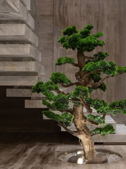 A low-angle shot emphasizing the grandeur of the faux bonsai against the wood and marble textures of the staircase. The lighting from below creates dramatic shadows and highlights the tree's form.
