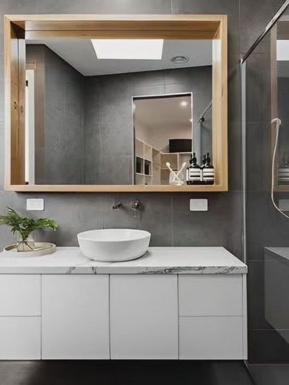 A contemporary bathroom with a floating white vanity and a large, wood-framed mirror. The walk-in shower with a glass panel and dark grey tiles completes the modern aesthetic.