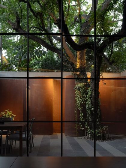 A view through a gridded window into the KEUS courtyard at night. The lighting highlights the texture of the Corten steel walls and the form of the mature tree, creating a dramatic and artistic composition.