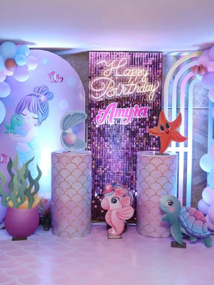 A vibrant and shimmering mermaid-themed backdrop. The combination of a purple sequin panel, a neon "Happy Birthday" sign, and pastel balloon arches creates a stunning focal point for the party.