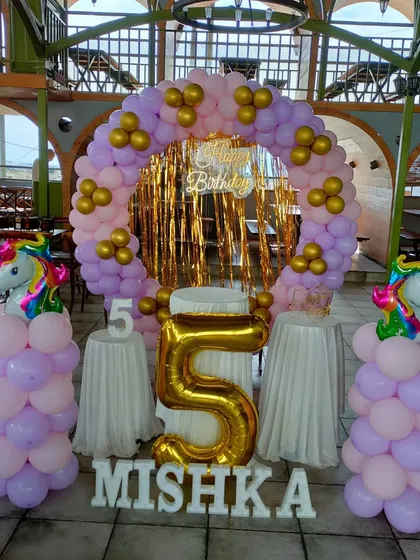A unicorn theme with a pastel purple and pink balloon ring against a gold tinsel backdrop. Unicorn-topped balloon pillars stand on either side.