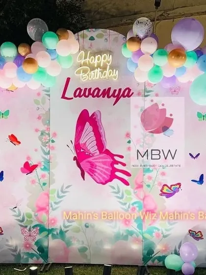 A lovely butterfly-themed backdrop with a large central butterfly graphic and a pastel balloon garland. The "Happy Birthday" neon sign adds a warm glow.