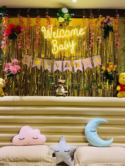 A cozy "Welcome Baby" decoration set up over a bed. It features a golden foil curtain, a neon sign, a name banner, and cute stuffed animals like Winnie the Pooh.