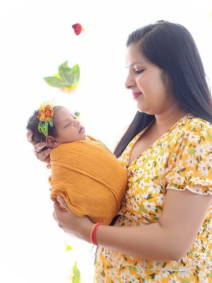 A bright and sunny newborn portrait. A new mother holds her baby, who is wrapped in a warm yellow swaddle, surrounded by flowers in our light-filled studio.