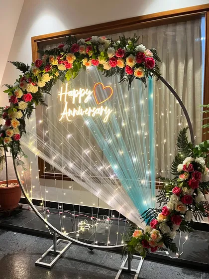 I design elegant backdrops like this anniversary ring for special celebrations. This setup features a large metal ring decorated with fresh roses and ferns, a custom 'Happy Anniversary' neon sign, and delicate fairy lights, creating a perfect photo opportunity.