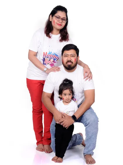 A full-length family portrait from a Holi photoshoot. The simple white outfits provide the perfect canvas for the bright colors, creating a classic festive memory.