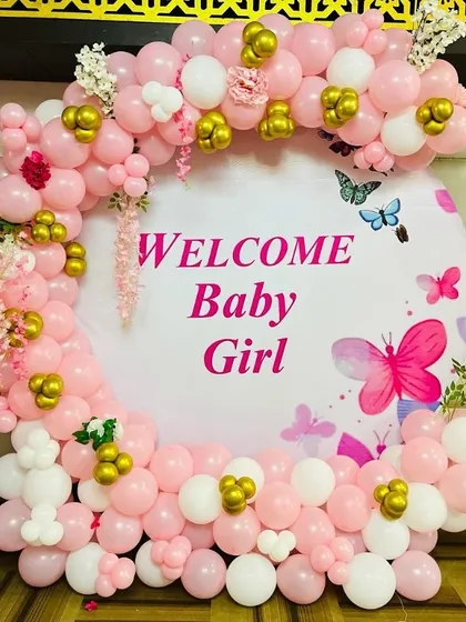 A beautiful "Welcome Baby Girl" backdrop. The decoration features a circular frame with a custom banner, surrounded by a pink, white, and gold balloon arch with butterfly accents.