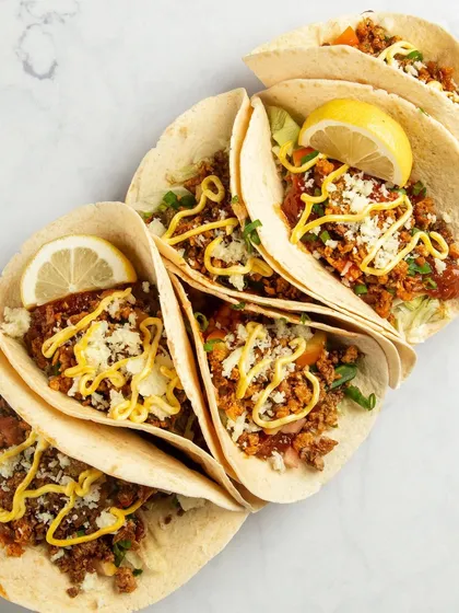 A top-down view of plant-based tacos. The composition creates a beautiful cascading pattern, and the bright toppings and lemon wedges make the dish look fresh and full of flavor.