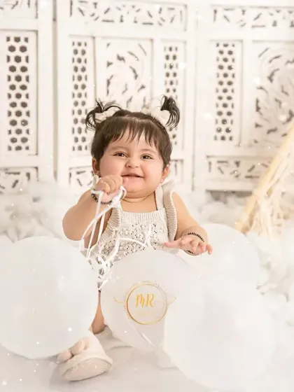 A baby girl in a boho-chic setup, playing with white balloons on a fluffy cloud-like rug.